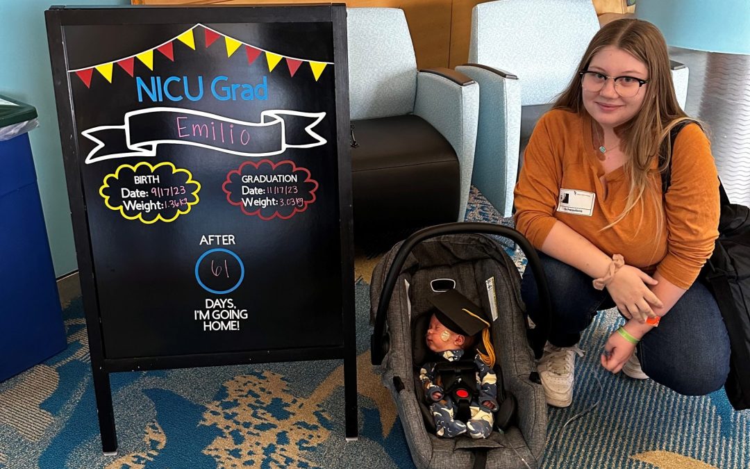 A tiny newborn baby in a carseat is in front of his mother who is kneeling beside a chalkboard. The Chalkboard reads, " NICU Grad Emilio. Birth Date, Weight 1.36 kg, Graduation Date, Weigh 3.03 kg. After 61 Days, I am going home!