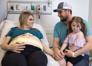 Mom wears monitor around her pregnant belly while holding her husband's knee as he rests their toddler girl on his knee