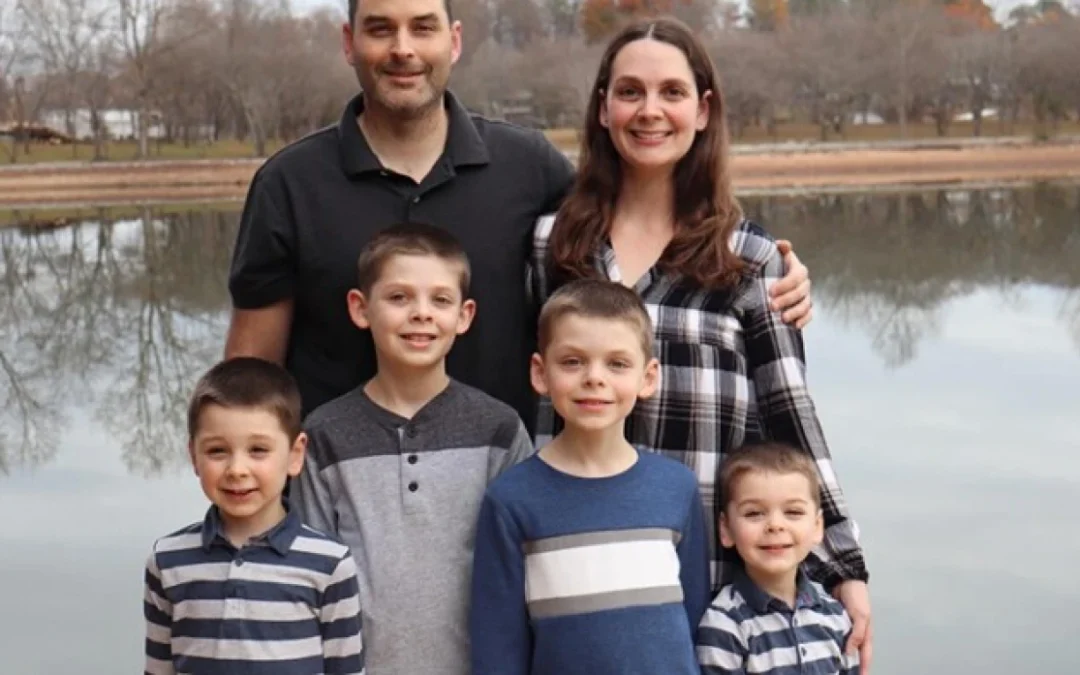 Mom, dad and four boys stand by a lake, smiling