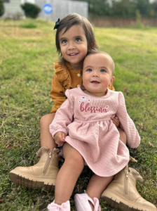 Smiling big sister holds baby sister who has recovered from a pericardial teratoma, looking up and looking happy