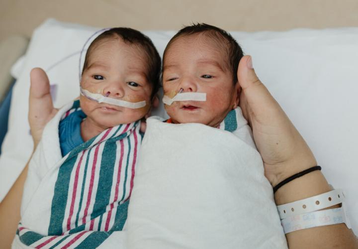 Twin baby boys lie side by side in the hands of their caregivers, both babies are wearing nasal tubes for oxygen supplementation