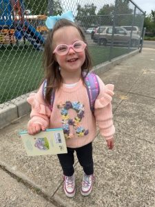 Little girl wearing a back pack and holding a book with a big smile walking to preschool 